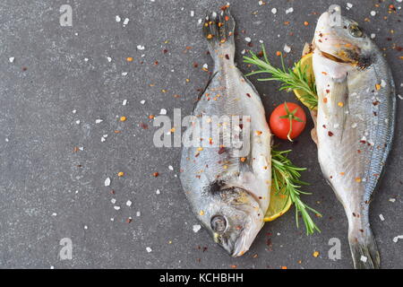 Frische rohe dorado Fisch auf einem Stück Papier mit Zitrone, Rosmarin, Zitrone, Cherry Tomaten auf grauem Hintergrund. gesunde Ernährung Konzept. mediterrane Leben Stockfoto