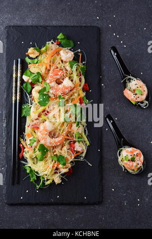 Nudeln mit Garnelen und Gemüse auf einem schwarzen Stein Platte und im traditionellen Suppe löffeln auf einem grauen Abstrakt Hintergrund. gesunde Ernährung Konzept Stockfoto