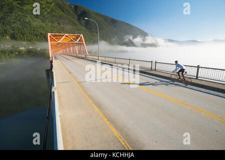 Ein Mann fährt mit dem Rennrad auf den Weg über die grosse orange Brücke in Nelson, BC Stockfoto