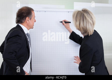 Reife Geschäftsfrau schreiben auf Flipchart während männliche Kollegen im Büro suchen Stockfoto