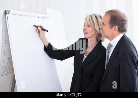 Reife Geschäftsfrau schreiben auf Flipchart während männliche Kollegen im Büro suchen Stockfoto