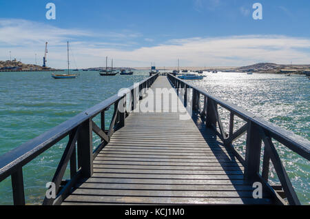 Hölzerne Seebrücke, die sich in der Bucht an einem sonnigen Tag und viele Boote, Lüderitz, Namibia, Südafrika Stockfoto