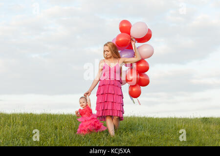 Mutter und Tochter in der schönen Kleider sind zu Fuß entlang der grünen Gras. Mama hält ein Bündel Luftballons in den Händen Stockfoto