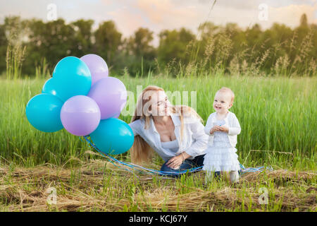 Junge Mutter und ihre Tochter sitzen auf dem Gras. Mama hält Ballons Stockfoto