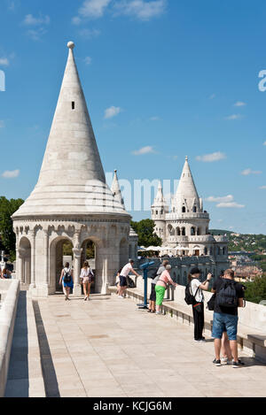 Touristen an den Wänden der Fischerbastei in Budapest betrachten die Aussicht und fotografieren Fotos an einem sonnigen Tag mit blauem Himmel. Stockfoto