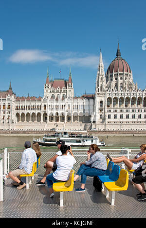 Touristen genießen eine Flussfahrt auf der Donau, während sie an einem sonnigen Tag mit blauem Himmel am ungarischen Parlamentsgebäude vorbeigeht. Stockfoto