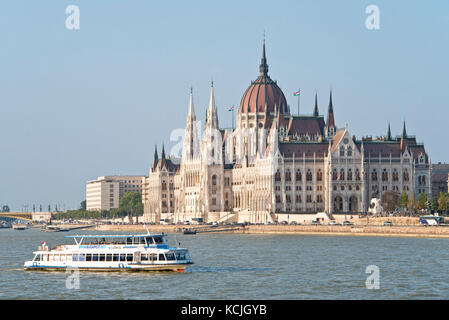 Ein Blick auf das ungarische Parlamentsgebäude an der Donau in Budapest mit einem Flussschiff, das an einem sonnigen Tag mit blauem Himmel vorbeifährt. Stockfoto