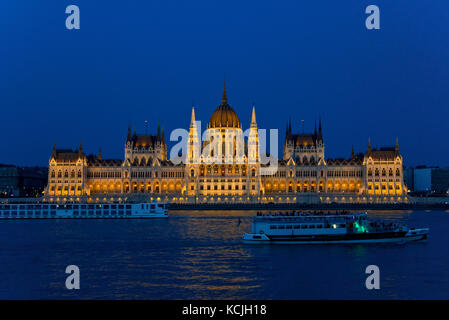 Ein abendlicher Blick auf das ungarische Parlamentsgebäude an der Donau in Budapest mit einem touristischen Flussschiff, das vorbeifährt. Stockfoto