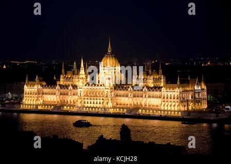 Eine Luftaufnahme von der Stadt am Abend mit Blick auf das ungarische Parlamentsgebäude in Budapest, an dem ein Touristenboot vorbeifährt. Stockfoto