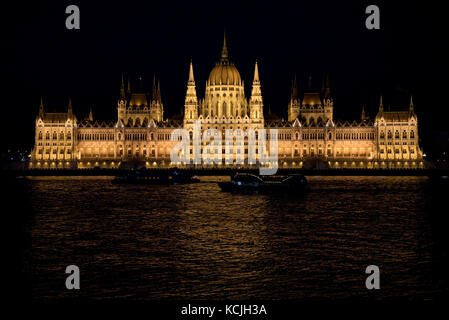 Ein abendlicher Blick auf das ungarische Parlamentsgebäude an der Donau in Budapest mit einem touristischen Flussschiff, das vorbeifährt. Stockfoto