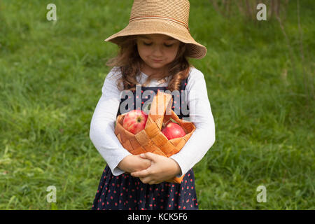 Verträumte kleine sechs Jahre altes Mädchen in wicker hat halten in ihren Händen Weidenkorb mit roten reife Äpfel im Garten hautnah. Stockfoto