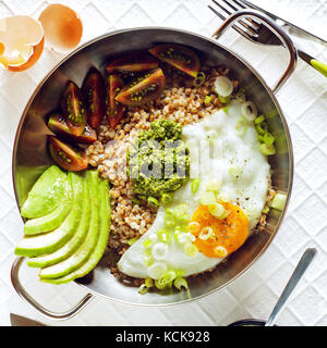 Frühstück mit Eiern, Avocado, Salat von Tomaten, Pesto und Buchweizen in einer Pfanne auf dem Tisch Stockfoto