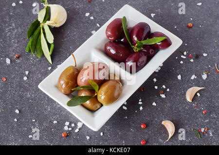 Schwarze und Grüne Oliven in eine weiße Platte auf einem grauen Abstrakt Hintergrund mit Zitrone, Knoblauch, Gewürze. Mediterrane Küche. gesunde Ernährung Konzept Stockfoto