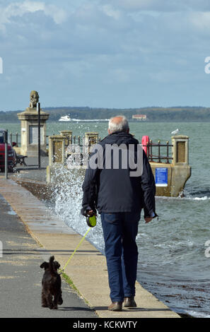 Ein Mann älter oder ältere einen kleinen schwarzen Hund an der Leine entlang der Strandpromenade an einem breezy Tag mit Spray brechen pver das Meer Wand wandern. Am Meer. Stockfoto