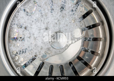 Weißer Schaum mit Blasen der cleane in einem Waschbecken. Ablassloch mit Seifenblasen in Metall Waschbecken, Makro anzeigen. Mechanisch einstellbare Ablassschraube Nahaufnahme. Stockfoto