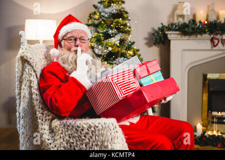 Weihnachtsmann mit Geschenk "Jobticket" sitzen auf Stuhl mit Finger auf die Lippen im Wohnzimmer während der Weihnachtszeit Stockfoto