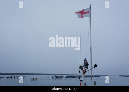 Die englische Flagge auf einem Fischerboot flattert auf eine schwache Brise, mit einem alten Hafen Steg und eine Rückkehr Boot vom Northumbrian Nordsee, am 25. September 2017, im Amble, Northumberland, England. Stockfoto