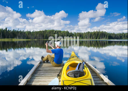 Mann sitzt auf dem Dock neben seinem Kajak bei Verwendung eines Tablet, 2 Mile Lake, Duck Mountain Provincial Park, Manitoba, Kanada Stockfoto