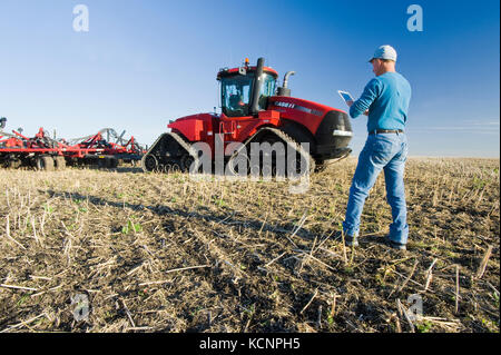 Landwirt mit einem Tablet vor einem Traktor und pneumatische Sämaschine Aussaat Winterweizen in einem Null bis Feld mit Raps Stoppeln, in der Nähe von Lorette, Manitoba, Kanada Stockfoto