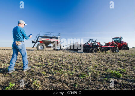 Landwirt mit einem Tablet vor einem Traktor und pneumatische Sämaschine Aussaat Winterweizen in einem Null bis Feld mit Raps Stoppeln, in der Nähe von Lorette, Manitoba, Kanada Stockfoto