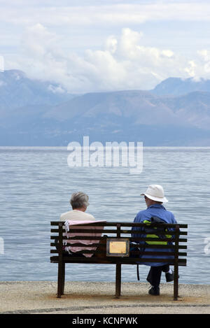 Ein älteres Paar auf einer Bank sitzen an der Küste mit Blick auf das Meer und die Berge. Stockfoto