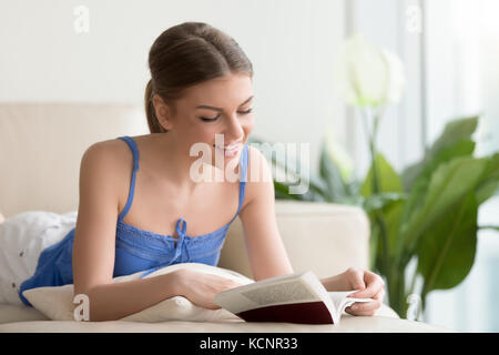 Junge lächelnde Frau liest interessante moderne Fiction Literatur auf der Couch zu Hause liegen, ziemlich Buch geliebte freie Zeit zu verbringen mit Bestseller, glücklich Stockfoto