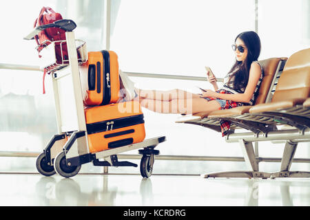 Jungen asiatischen reisenden Frau, Student, sitzen Sie ihr Smartphone am Flughafen, Gepäck und Tasche auf Trolley Warenkorb. online Check-in mobile App Konzept Stockfoto