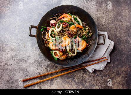 Buchweizen unter Rühren braten, Nudeln mit Meeresfrüchten - Garnelen, Tintenfisch, Tintenfisch in Gusseisen Asiatischen Wok mit Essstäbchen. Ansicht von oben, Stein Hintergrund. Stockfoto