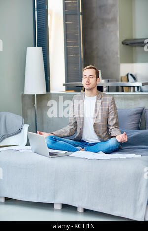 Ein junger Mitarbeiter im Home Office sitzt in Lotus-Pose auf seinem Bett, um sich zu entspannen Stockfoto