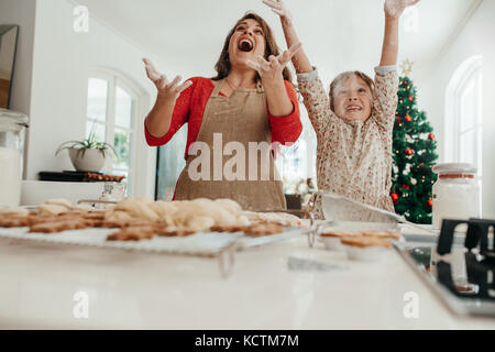 Glückliche Mutter und Tochter in der Küche werfen Mehl in die Luft, während die Cookies. Gebackene Kekse, Muffins und Lebkuchen am Küchentisch mit Dekorieren Stockfoto
