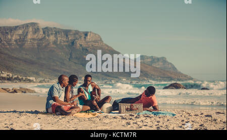 Afrikanischer Mann Gitarre spielen für seine Freunde entlang der Küste. Gruppe von Menschen Entspannung am Sandstrand. Stockfoto