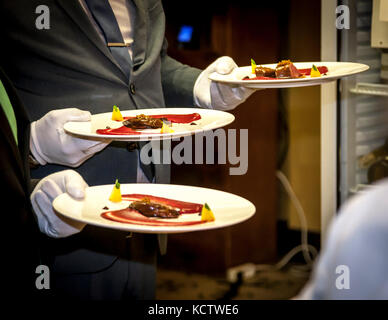 Ein Kellner in einem grauen Anzug und weißen Handschuhen serviert drei Teller mit einem Gourmet-Gericht aus dunkel glasiertem Fleisch, roten Püreebögen und hellgelben Würfeln mit kleinen Kräuterblättern im Hotel und Restaurant Kronenschlösschen in Hattenheim, Eltville am Rhein, Hessen, Deutschland. Ein Kollege in einem dunklen Anzug steht links. Das Gericht ist Teil des Galadinners, das der mit einem Michelin-Stern ausgezeichnete Koch Thomas Kellermann am Abend des 8. März 2017 im Kronenschlösschen zubereitet hat. Das Dinner ist Teil des 21. Rheingau Gourmet & Wein Festivals. Es umfasst fünf Gänge. Caro Maurer, Master of Wine, moderiert den Abend. Stockfoto