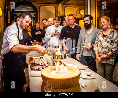 Gäste auf der Küchenparty warten auf ein flambiertes Risotto im Hotel und Restaurant Kronenschlösschen in Hattenheim, Eltville am Rhein, Hessen, Deutschland. Flammen lodern im ausgehöhlten Rad des Parmigiano Reggiano (Parmesan) und in der Pfanne, die ein junger Koch darüber hält. Die Gäste stehen am Tisch, und ein Mann macht mit seinem Smartphone ein Foto. Neben ihm steht ein bärtiger Mann mit Brille, der ein Weinglas hält, und eine ältere Frau mit gefalteten Händen. Am Abend des 12. März 2017 endet das 21. Rheingau Gourmet & Wein Festival mit der Abschiedsparty "Burgenland". Das Kronenschlösschen feier Stockfoto