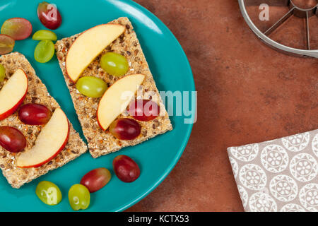 Vergoldete multigrain Knäckebrot Schichten mit Apple Segmenten und Traube Hälften auf einem Naturstein Arbeitsfläche gekrönt Stockfoto