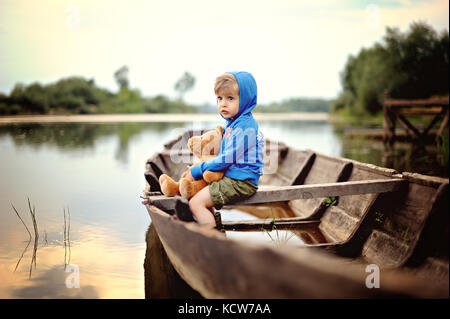 Eine kleine, nachdenkliche Junge in einem blauen Kapuzenshirt sitzt auf bewaldeten Boot mit Teddybär Stockfoto