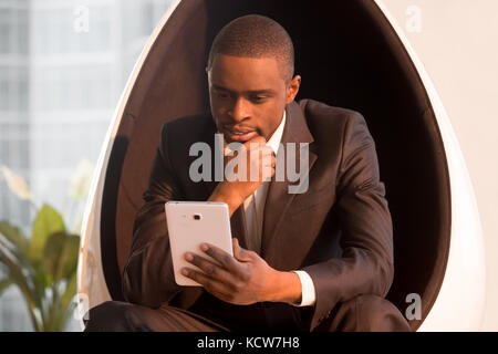 Porträt eines erfolgreichen nachdenklichen afroamerikanischen Geschäftsmannes im stilvollen Anzug, der auf futuristischem Eierstuhl sitzt und ein digitales Tablet verwendet. Schönes schwarzes Modell Stockfoto