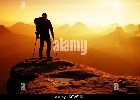 Touristische mit Medizin Krücke über den Kopf erreicht Berggipfel. Wanderer mit gebrochenen Bein in der Wegfahrsperre. tief nebligen Tal unten Silhouette der Mann mit Stockfoto