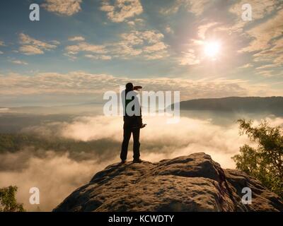 Professionelle Fotografen mit Stativ in den Händen von Cliff und Denken. verträumte fogy Landschaft, blauen Nebel Sonnenaufgang in einem schönen Tal, unten Stockfoto