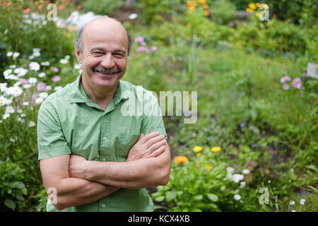 Ein älterer Mann mit einem Schnurrbart und eine kahle Stelle in ein grünes T-Shirt steht unter den Blumen im Sommer Garten, mit verschränkten Armen und lächelnd. Stockfoto