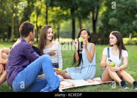 Freunde auf Picknick, junge Mann wirft einen Apfel zu Frau. Studenten Spaß am Mittagessen im sonnigen Park. Menschen unterschiedlicher Nationalitäten verbringen Ihre fr Stockfoto