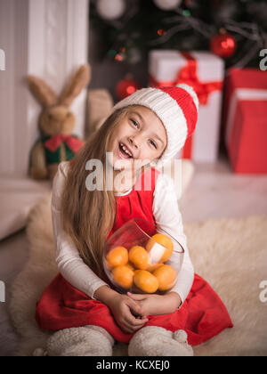 Schöne Mädchen in santa Kappe mit Mandarinen in der Hand, saß auf dem Boden im roten Kleid, Studio gedreht. Stockfoto