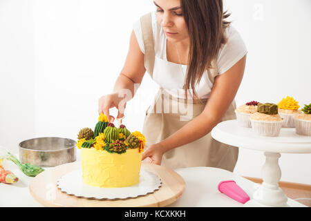 Eine attraktive junge Frau ein konditor in ein weißes T-Shirt, beige Schürze ist geschmückt mit einem gelben Diät Kuchen, dekoriert mit einer grünen Bohne Creme in einer Stockfoto