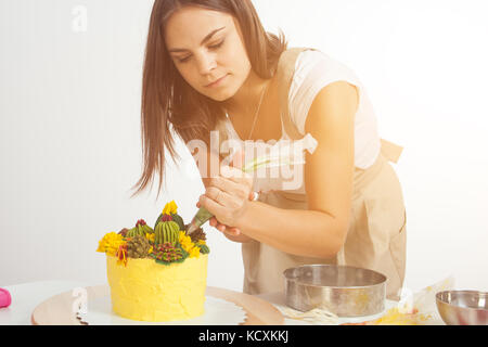 Eine dunkelhaarige Junge Frau in einem weißen T-Shirt, beige Schürze, schmückt einen Kuchen mit Gebäck Spritze, eine gelbe Diät Kuchen, mit einer grünen Bohne eingerichtet. c Stockfoto