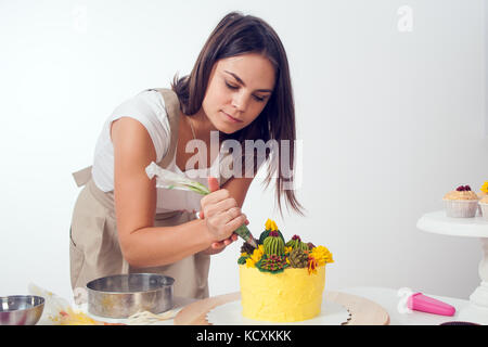 Eine dunkelhaarige Junge Frau in einem weißen T-Shirt, beige Schürze, schmückt einen Kuchen mit Gebäck Spritze, eine gelbe Diät Kuchen, mit einer grünen Bohne eingerichtet. c Stockfoto