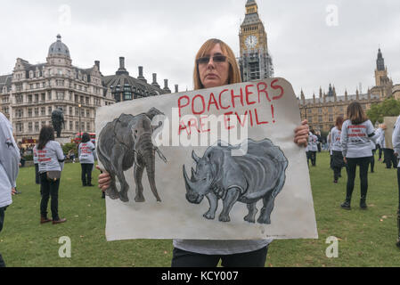 London, Großbritannien. 7. Oktober 2017. Eine Frau hält ein Plakat mit Wilderer die Meldung 'sind böse!' in der Diamond Bildung von Menschen in T-Shirts gekleidet, gegen den Handel mit Elfenbein mit einigen Holding mock Elephant tusks oder Rhino Hörner in eine halbe Stunde lang stillen Protest im Parlament Platz als Ereignis in die jährliche globale März für Elefanten und Nashörner (GMFER) rund um die Welt. Lautsprecher am Ende der Mahnwache auf der britischen Regierung rief mit einem vollständigen Verbot auf Elfenbein zu bringen und am Ende der Handel mit Antiquitäten Händler in Großbritannien, spielt eine wichtige Rolle in Côte d'Ivoire, die Märkte offen zu halten und dem Fahren des Pochieren