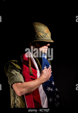 Patriotischer amerikanischer Soldat (US Marine / Vietnam Krieg) mit der Stern- und Sripes-Flagge um seine Schultern, vor dunklem Hintergrund. Stockfoto