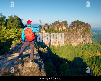 Touristische mit roten Rucksack nimmt Fotos mit Smart Phone von Felsen und Tal. im Frühling oder im Sommer bei schönem Wetter, frische grüne Farben Stockfoto