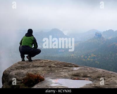 Touristische in Rot und Grün Schwarz Sportswear in hockender Stellung auf einem Felsen, Herbst Landschaft genießen. lange Tal nach regnerischen Voller schwere cremige Nebel Stockfoto