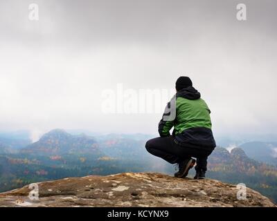 Touristische in Rot und Grün Schwarz Sportswear in hockender Stellung auf einem Felsen, Herbst Landschaft genießen. lange Tal nach regnerischen Voller schwere cremige Nebel Stockfoto