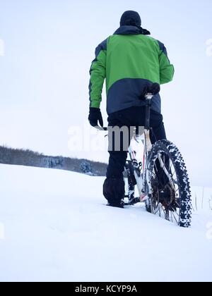 Mann mit Mountainbike Aufenthalt im Pulverschnee. verloren weg in tiefe Schneeverwehungen. Hinterrad detail. Schneeflocken schmelzen in dunklen off road Reifen. Winter Wetter können Sie Stockfoto